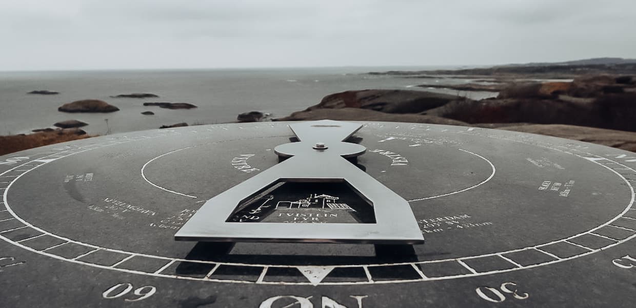 Stort kompass-monument på Rakke med utsikt over havet. Den metalliske kompassnålen peker mot nord over en sirkulær steinplate med kompassrose og graderinger. I bakgrunnen ses svaberg og hav i en gråværsdag.